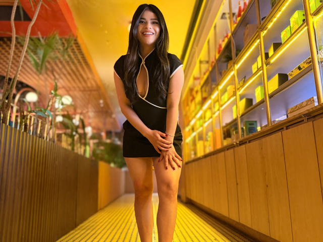PaulinaValley standing in a yellow-lit hallway, smiling, wearing a black dress with a deep cleavage, hands near her thighs, long dark hair, looking cute, showing her ass.