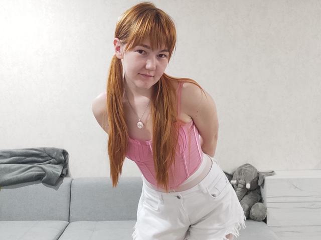 SheritaChieng mit roten Zöpfen, einem pinken Tanktop, weißen Hosen und einer Halskette, die nach vorne gebeugt, lächelnd, mit kleinen Brüsten, süß und schlank, auf einem grauen Sofa..