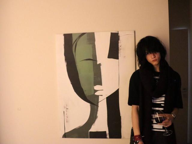 Een foto toont LegacyStar met rommelig zwart haar, gekleed in een zwarte trui en sjaal, die rechts staat van een abstract schilderij in groen en zwart. Ze houdt een glas vast..