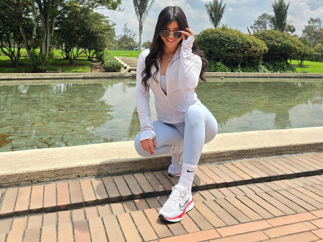 PaulinaValley squatting, wearing white jacket, blue leggings, white sneakers, red Nike logo, and red socks, adjusting sunglasses, with long dark hair, in a sunny park.