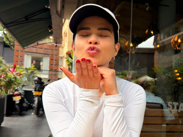 PaulinaValley with pouty lips, red nails, and white long-sleeve shirt, hands near face, black cap, brown skin, and cute expression, sitting outside, blurred background.