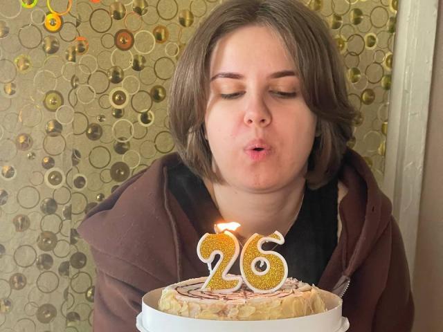 Mary Cordy mit kurzen, braunen Haaren, vollen Lippen, die einen braunen Kapuzenpullover trägt und eine goldene Zahl 26 auf einer verzierten Torte ausbläst..