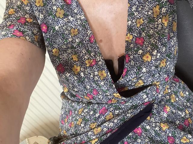Bourgeoisex's floral dress has a deep V-neck, exposing a black bra and her bare chest. Her skin is light with a small mole. She's seated with a black strap.