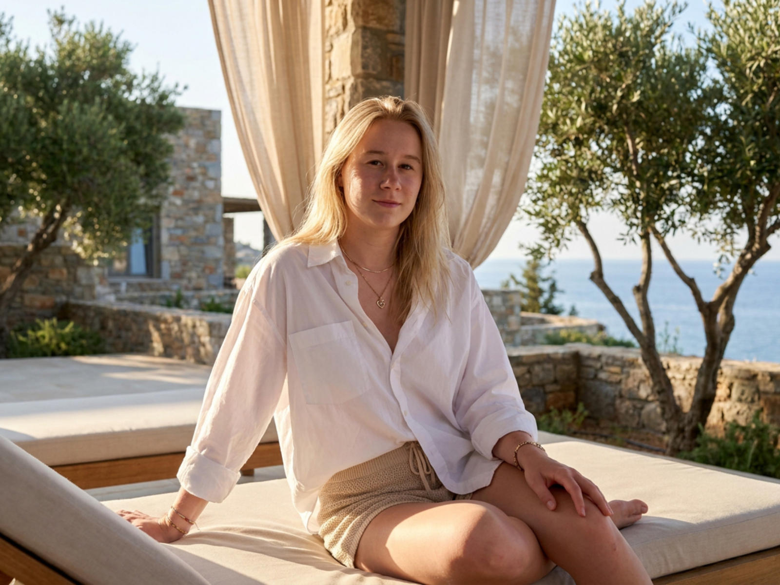 Leisa Farrill mit blonden Haaren, einem weißen Hemd und beigen Shorts, sitzt auf einem sonnigen Liegebett im Freien, ihr Hintern ist leicht sichtbar, sie wirkt entspannt und hat einen ruhigen Gesichtsausdruck..
