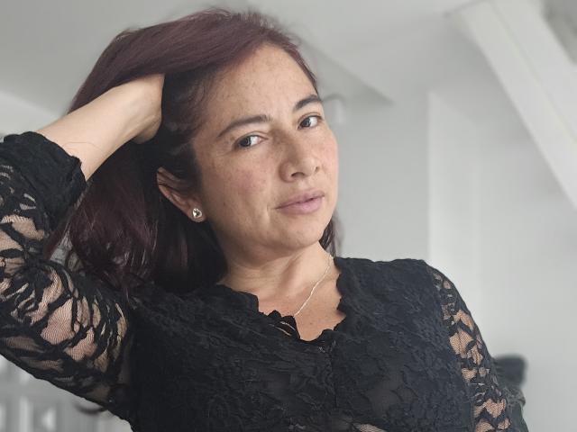 Alessia, pele marrom média e cabelo ruivo, vestindo uma blusa preta de renda, com a mão no cabelo, olhando para a câmera, pequenos brincos de diamante, sorriso discreto, maquiagem suave..