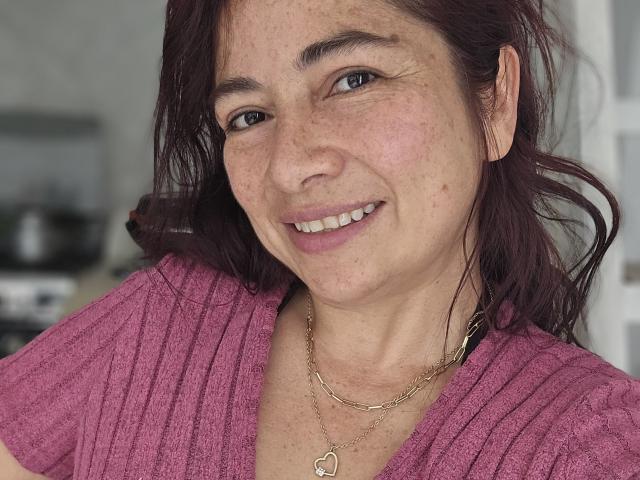 Alessia: Rotes Haar, Sommersprossen im Gesicht, rosafarbenes Rippstrickshirt, mittelbraunes Haar, leicht lächelnd, trägt eine goldene Herz-Kette und eine goldene Kette, in einem ungezwungenen Selfie..