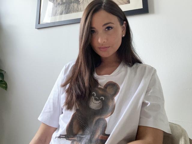 Dionisia with long brown hair, wearing a white tee with a bear graphic, sitting, looking at camera, subtle smile, small earrings, light skin, cute, casual, relaxed.
