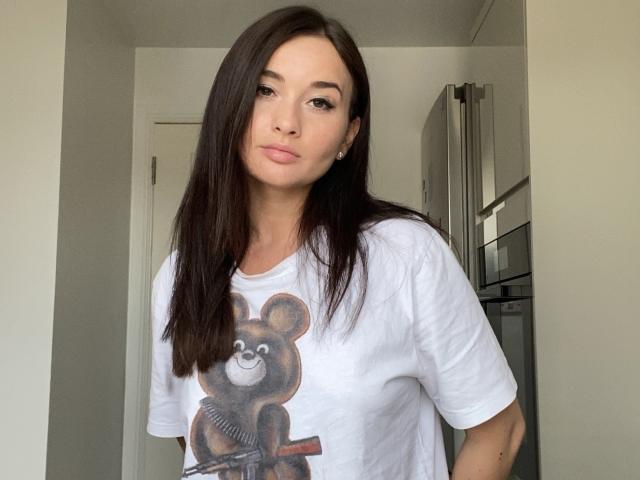 Dionisia with long dark brown hair, wearing a white tee with a bear graphic, standing in a kitchen, looking at the camera with a slight pout, tits visible.