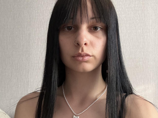 VirginiaAttaway with long black hair, nose ring, and pearl necklace, staring at the camera with a serious expression, showing her bare shoulders and soft skin.