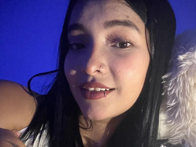 SaritaKlart with black hair, nose ring, and lip piercing, smiling with slightly parted red lips, blue background, and white fluffy pillow on the right.
