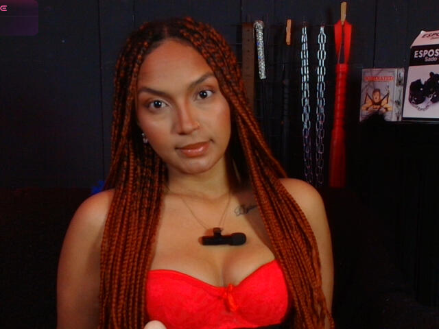 BrianaPratt with long orange braids, red lace bra showing her tits, black necklace with a key, tattoo on right side of neck, dark background, serious expression.