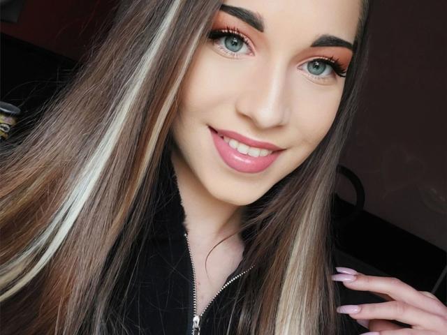 Chatgirl atractiva, con cabello castaño largo, labios rosados, ojos verdes y una chaqueta negra, sonriendo a la cámara, mostrando uñas rosadas y un rostro sexy y lindo..