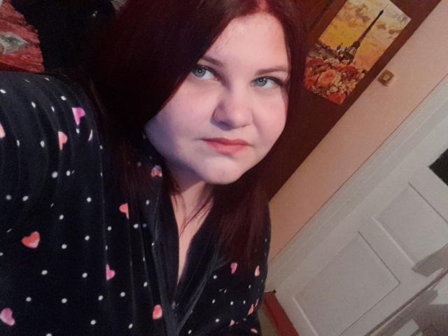 PollyEmm, avec des cheveux roux et des yeux bleus, portant une robe noire à pois avec des cœurs roses, regardant la caméra avec une expression neutre..