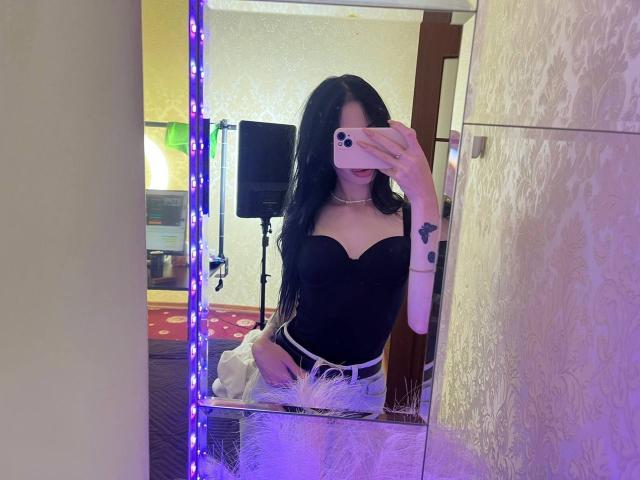 SamantaBredShow with long black hair, wearing a black strapless top, white belt, and holding a phone, reflecting in a mirror with purple LED lights. Visible tattoo on right arm.
