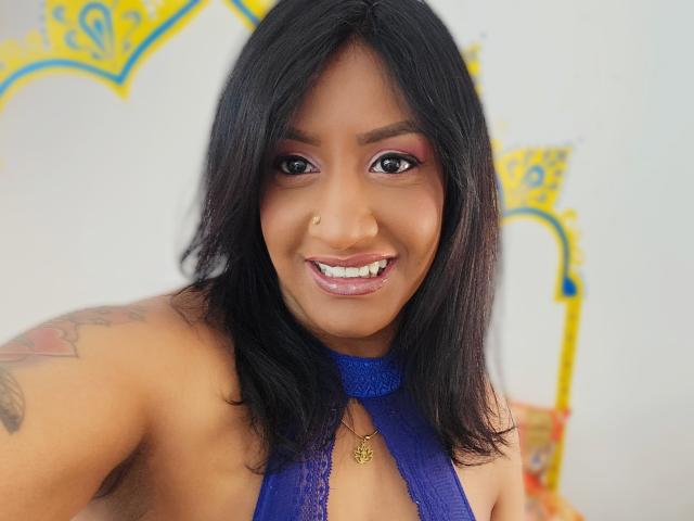 Nahirr ha una pelle marrone media, capelli neri lunghi fino alle spalle, trucco viola per gli occhi, un top con spalline sottili color blu, un tatuaggio visibile sulla spalla destra e una collana d'oro, sorridendo alla fotocamera..
