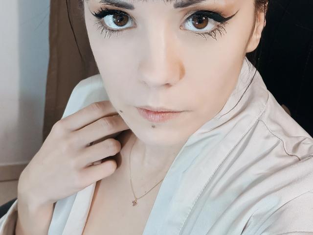 OrianaLaFrancaise with pale skin, brown eyes, and winged eyeliner, wearing a white shirt, has a small beauty mark below her right eye and a delicate necklace. Her hand rests near her neck.
