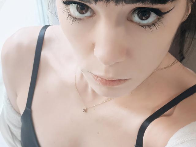 OrianaLaFrancaise's face with big brown eyes, thick black eyeliner, pale skin, small pink lips, black spaghetti strap top, gold necklace, and a slight off-shoulder white garment.