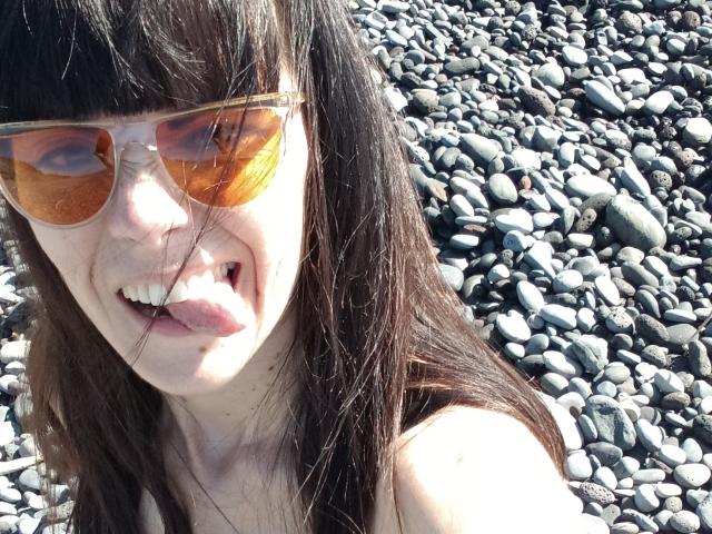 OrianaLaFrancaise with long brown hair, sticking out her tongue, wearing orange-tinted sunglasses, and showing her bare shoulders, standing on a rocky beach.