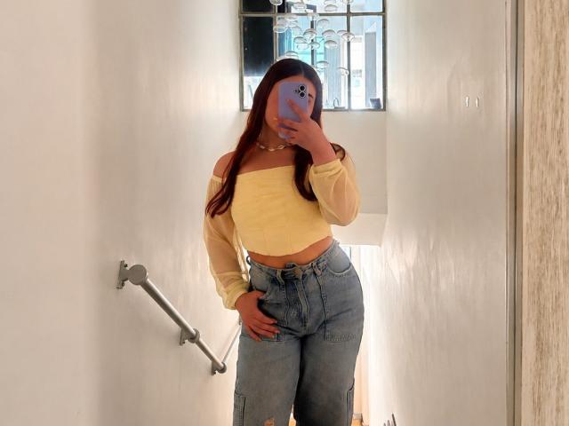VicluX mit langen, roten Haaren, einem gelben Top mit einem Schulterauschnitt und blauen Jeans mit hoher Taille, hält ein violettes Telefon und steht in einem weißen Flur, wobei ihr wohlgeformter Hintern zu sehen ist..
