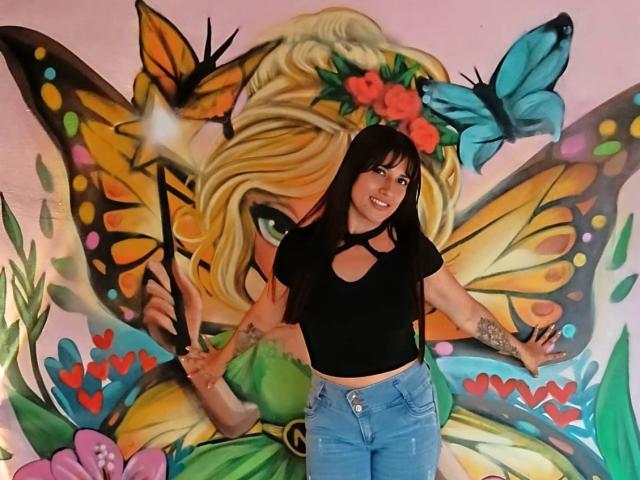 AlisRoberts has long brown hair, black top with cutouts, blue jeans, and tattoos on her arms. She's standing in front of colorful butterfly and flower mural with a blue butterfly.