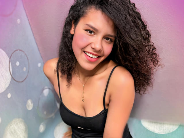 AriaVega with curly black hair, smiling, wearing a black spaghetti-strap top, showing her small tits and light brown skin, against a colorful, bubbly background.