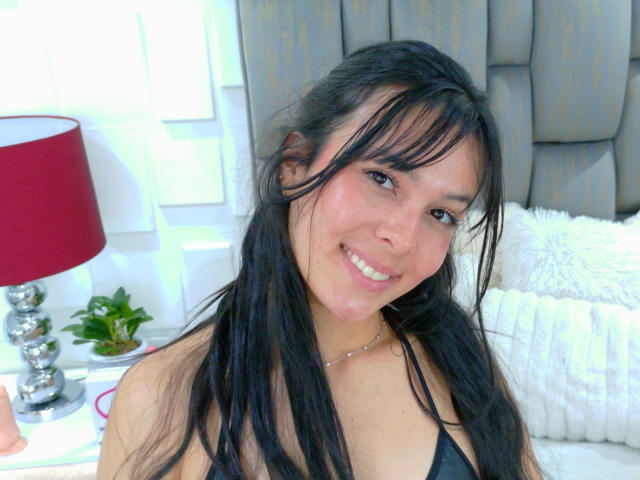 ValerieNoiir has long black hair, brown eyes, and a cute smile. She's wearing a black top and a silver necklace, with a red lamp and white pillow in the background.