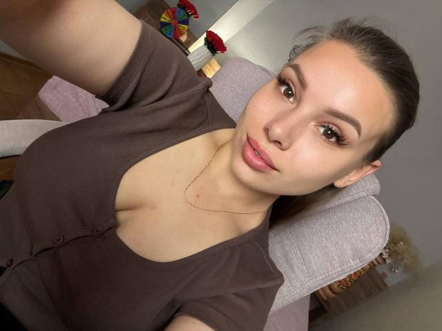 Niferttiti with fair skin, brown eyes, and pink lips, wearing a brown cardigan, showing her boobs, in a selfie, holding a phone, with a neutral expression.