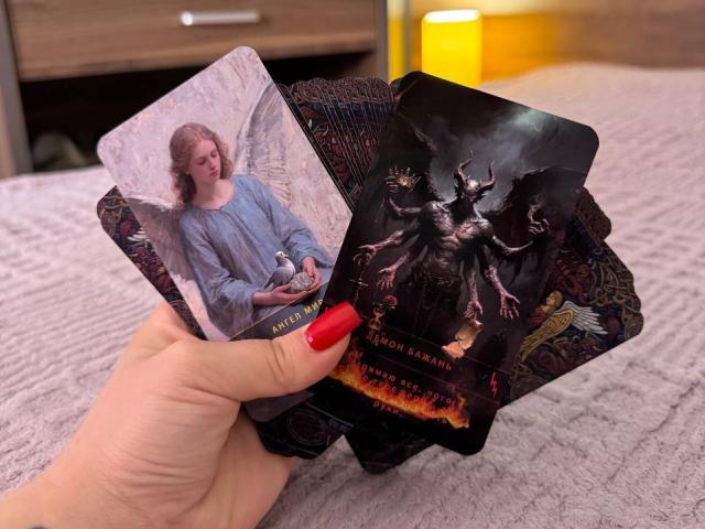 Hand holding two cards, Niferttiti's angelic card with white wings and blue dress, and dark demonic "The Book of Blood" card with red fire and black creatures. Niferttiti's red nails.