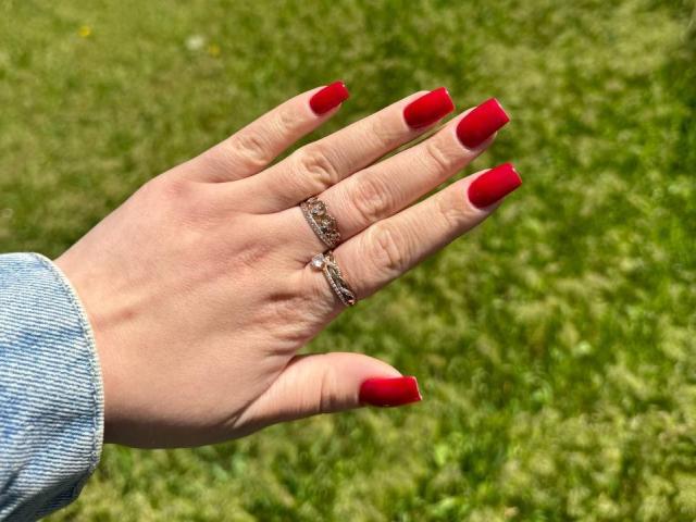 Niferttiti's hand with red nails, wearing two silver rings, and a blue denim sleeve, against a green grass background.