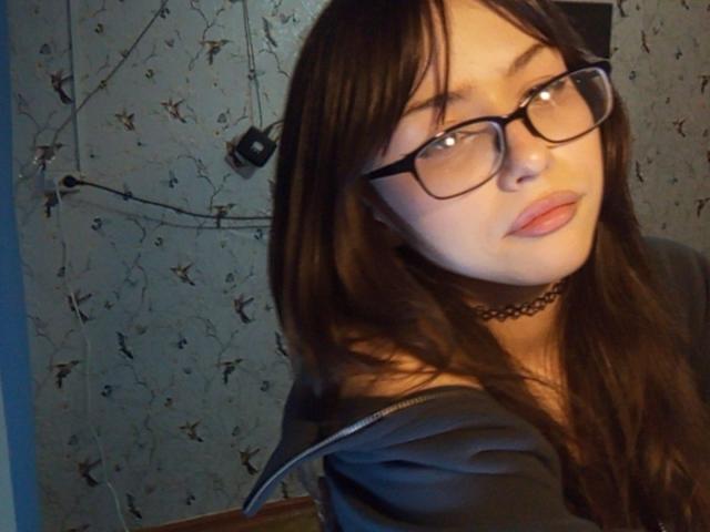 EileneSzala with long brown hair, black glasses, and a black choker, wearing an off-shoulder gray top, looking at the camera with a slightly pouty expression.