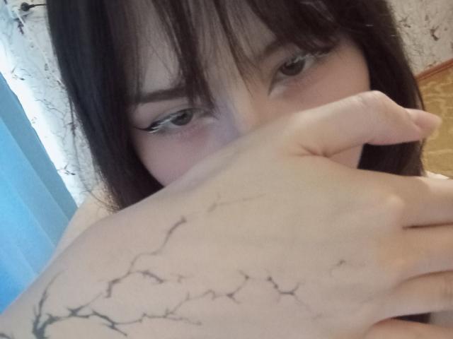 EileneSzala with dark brown hair, half-covered face, blue eyes, and a hand over her mouth, showing a black tree tattoo on her arm. Her nose is slightly pink.