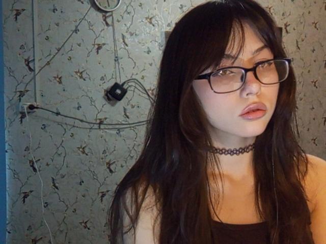 EileneSzala with long dark hair, glasses, choker, and pink lips, wearing a black top, looking at camera with slightly parted lips, in a dimly lit room.