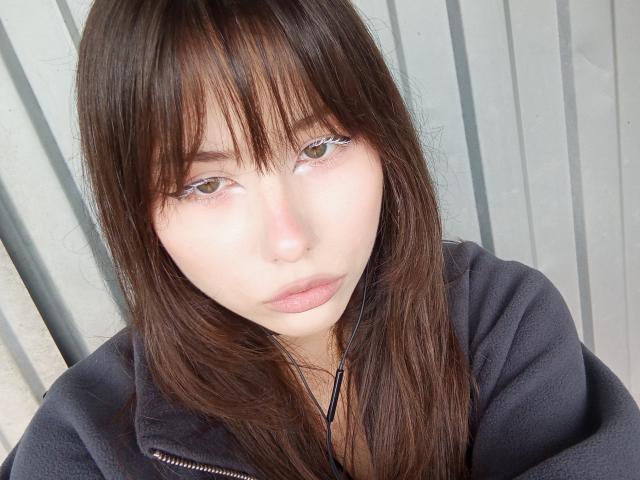 EileneSzala with long brown hair, bangs, and pale skin, wearing a black hoodie, looking straight into the camera with subtle pink lips and small nose, brown eyes.