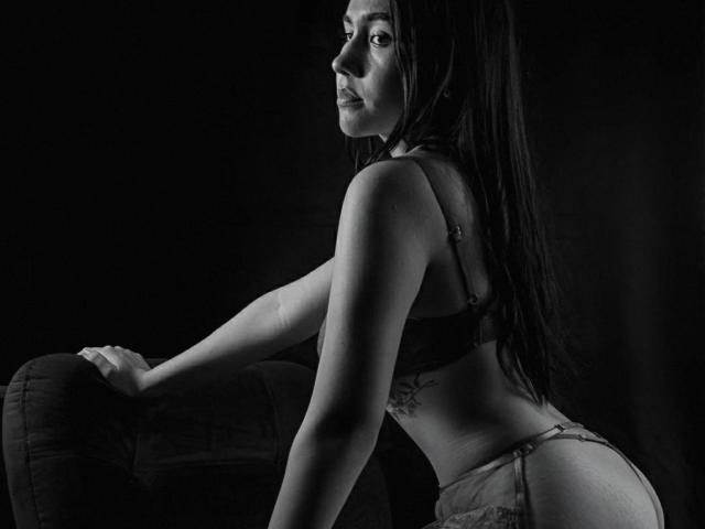 BibiSugarX with long dark hair, wearing a bra and thong, leaning on a chair in a black-and-white photo, showing her ass and looking over her shoulder.
