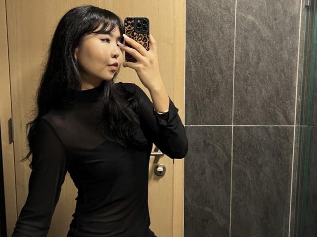 IrisDommer, com cabelo preto, vestindo uma blusa preta transparente de manga comprida, tirando uma selfie no espelho com uma capa de celular estampada com onças, mostrando seu rosto e seios pequenos..