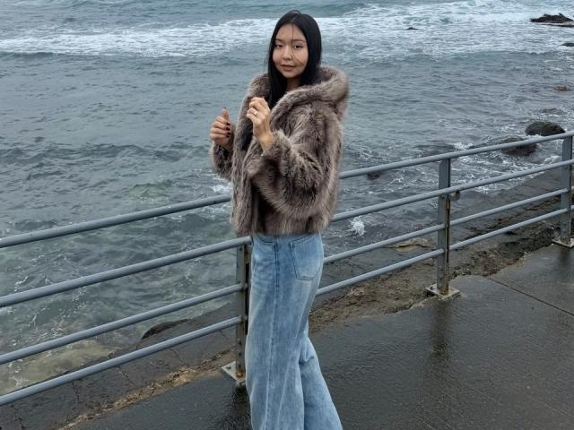 IrisDommer mit langem, schwarzem Haar, die eine flauschige, braune Felljacke und hoch taillierte, blaue Samthosen trägt, steht an einem Metallgeländer am Meer, die Hände in der Nähe ihrer Brust..