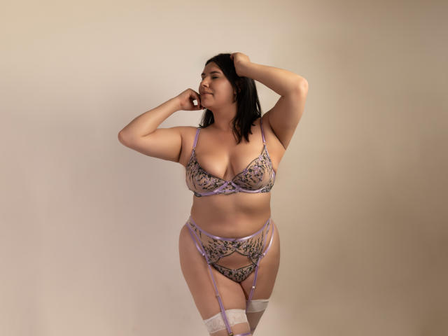 ZoeyGrey, avec une peau pâle, des cheveux noirs, portant un soutien-gorge en dentelle violet, une jarretière assortie et des bas blancs montants jusqu'à la cuisse, une main dans les cheveux, debout, montrant ses seins et ses fesses..