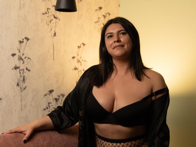 ZoeyGrey com sutiã preto, meia-calça de rede e robe preto aberto, mostrando os seios, sentada em uma cadeira rosa, sorrindo para a câmera, sob um abajur preto..