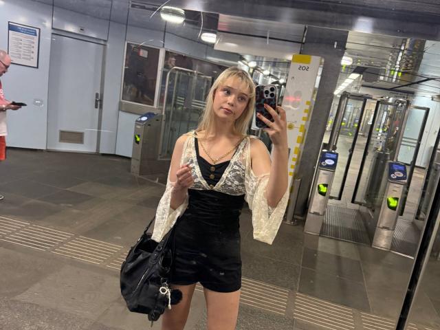 DonitaDunn with blonde hair, wearing a black lace-up top and sheer white sleeves, taking a mirror selfie in a subway, holding a black bag, showing her tits.