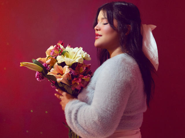 Avril Thomas hält einen Strauß aus rosa, gelben und orangefarbenen Blumen, trägt einen flauschigen grauen Pullover und eine weiße Schleife in ihrem dunklen Haar und lächelt sanft..