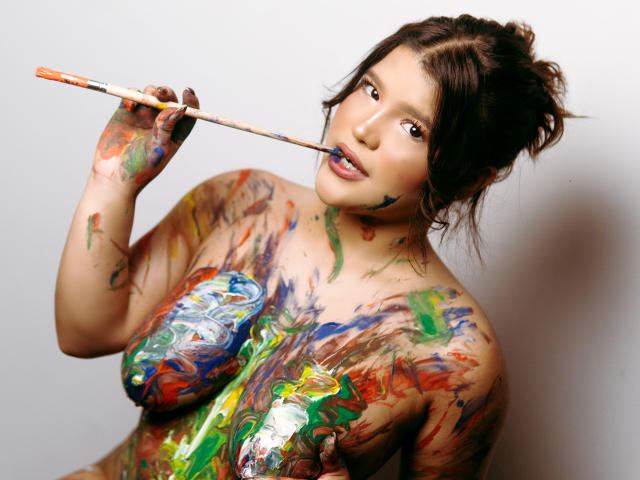 Avril Thomass, coberta de tinta colorida, segura um pincel perto dos lábios pintados de azul, com tinta nos seios, nádegas e mãos, olhando sedutivamente para a câmera..