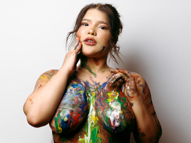 Avril Thomass ha della vernice colorata su seni, glutei e viso, con le mani che toccano le labbra e il petto dipinti, in piedi su uno sfondo bianco..