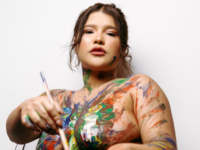 Avril Thomass, con salpicaduras de colores en el pecho y los brazos, sostiene un pincel en su mano derecha, mirando a la cámara con una expresión neutra..