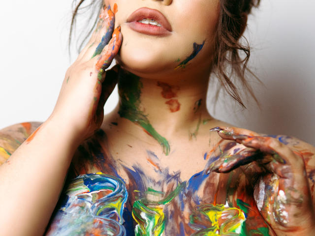 Le visage et la poitrine d'Avril Thomass sont couverts de peinture colorée, et ses mains touchent son visage et sa poitrine. Ses lèvres sont légèrement entrouvertes, laissant entrevoir un sourire subtil..