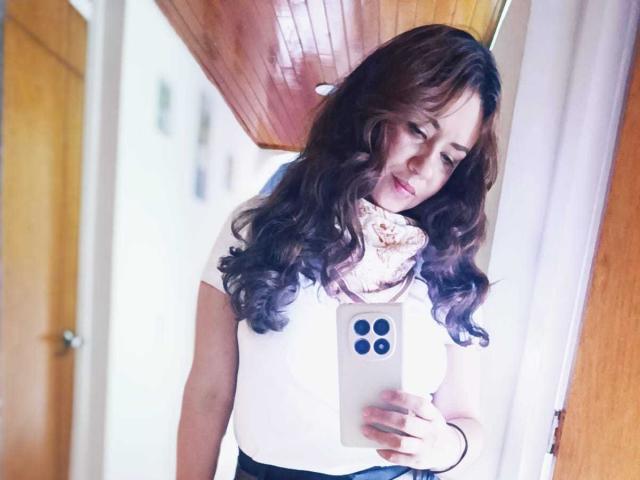 TheresaLarson with wavy brown hair, white shirt, pink scarf, black skirt, holding a white iPhone, looking down, wooden ceiling, wooden door, white walls, soft light.