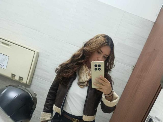 TheresaLarson with wavy brown hair, wearing a brown leather jacket, white shirt, and beige scarf, taking a mirror selfie with a beige phone, showing her tits slightly.