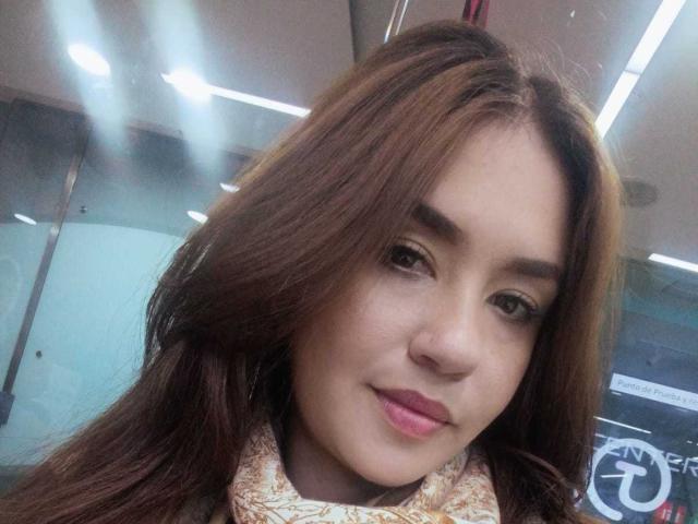 TheresaLarson's face, with long brown hair, brown eyes, and pink lipstick, showing a slight smile. She's wearing a patterned scarf, looking cute and attractive.
