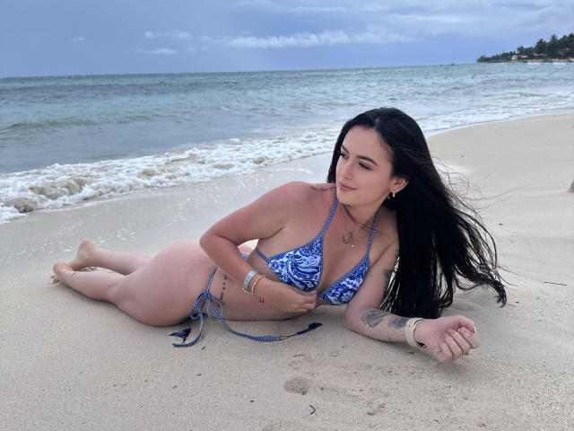 SofiaLopezHot lies on a sandy beach, wearing a blue and white patterned bikini, covering her tits with one hand, her black hair flowing, tattoos on her arm.