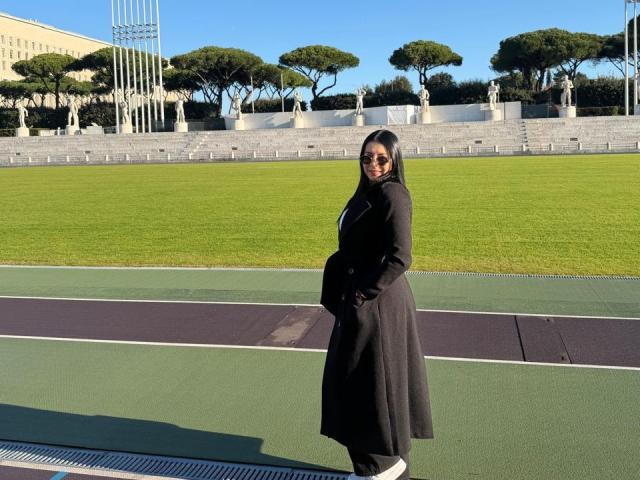 IsabellaJames, curvy with a big ass, wearing a black long coat and sunglasses, stands on a green and purple sports field, hands in pockets, looking over her shoulder.