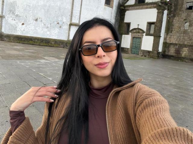 IsabellaJames with long black hair, brown eyes, wearing sunglasses, a brown rib-knit jacket, and a maroon turtleneck, pouting her pink lips, touching her hair with a manicured hand.